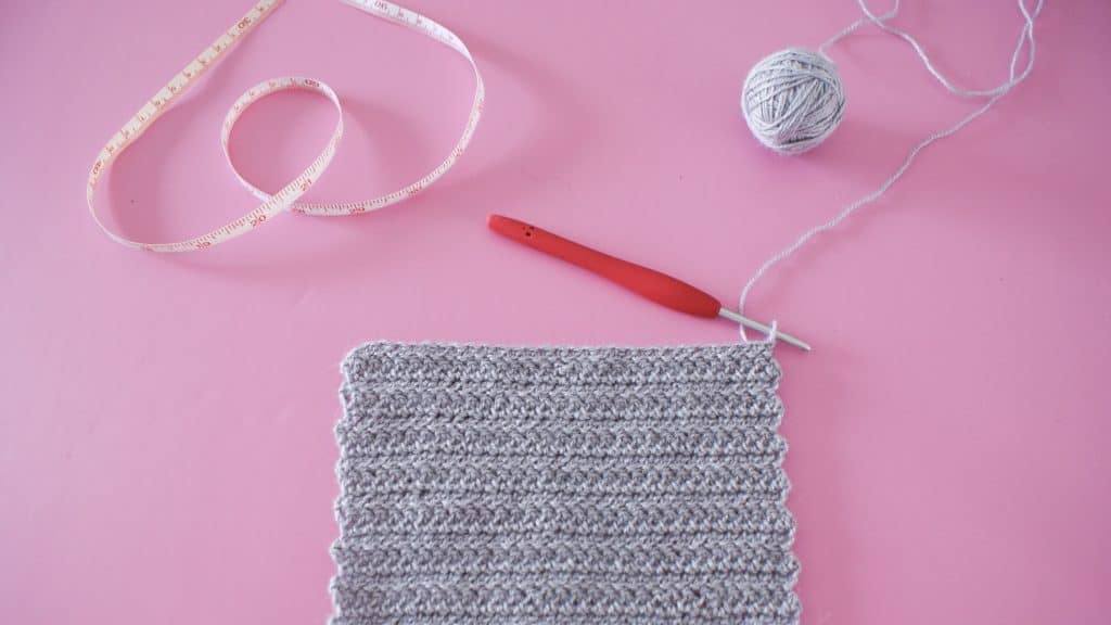 Grey herringbone crochet swatch on pink background with red hook, ball of yarn and tape measure