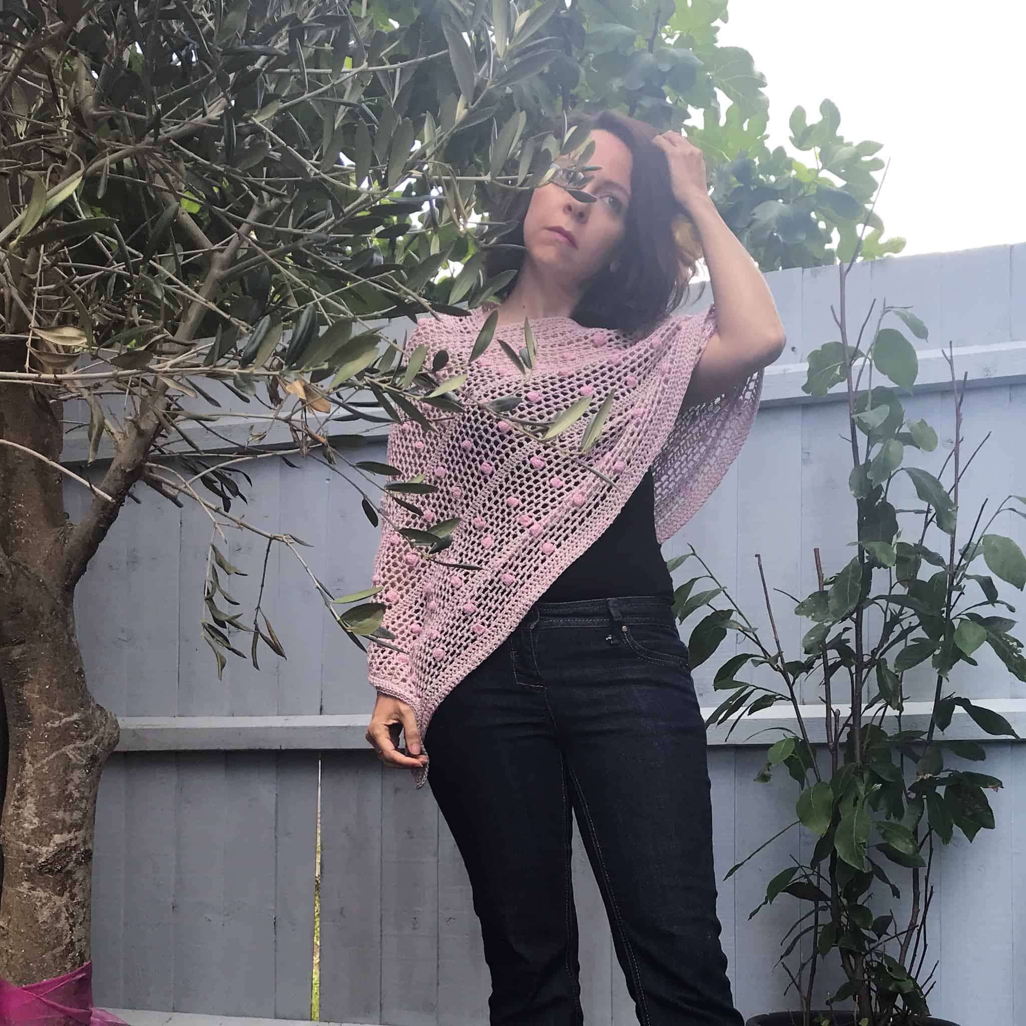 Woman standing in front of blue fence amid trees wearing pink and green crochet poncho