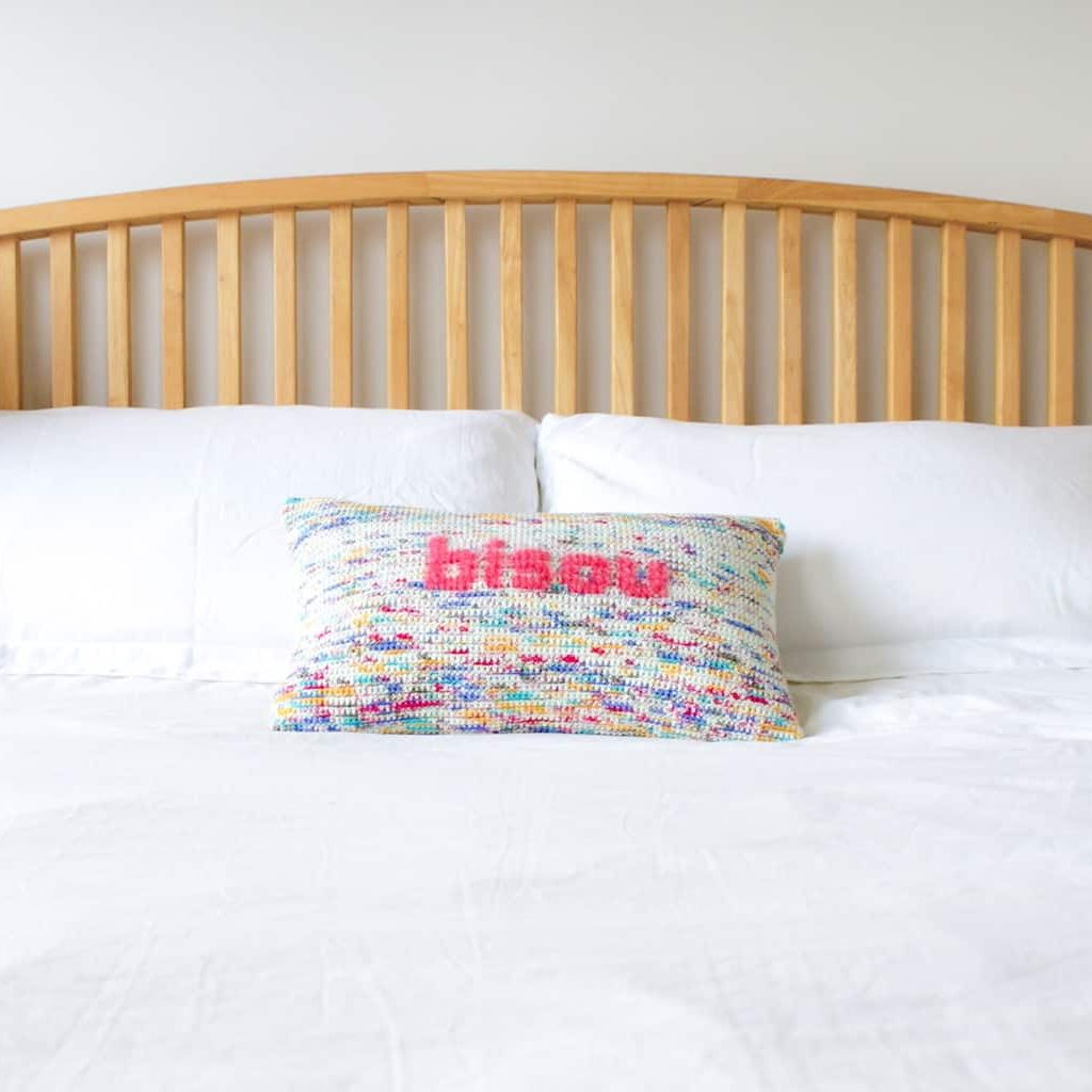 Multi colour crochet pillow with pink fluffy lettering reading bisou sits on a bed with white linen