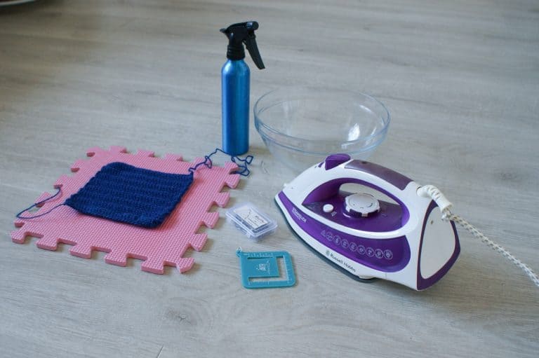 A collection of materials ready to block a crochet swatch including an iron, water bowl, water spray, blocking board, pins, swatch measure and a swatch