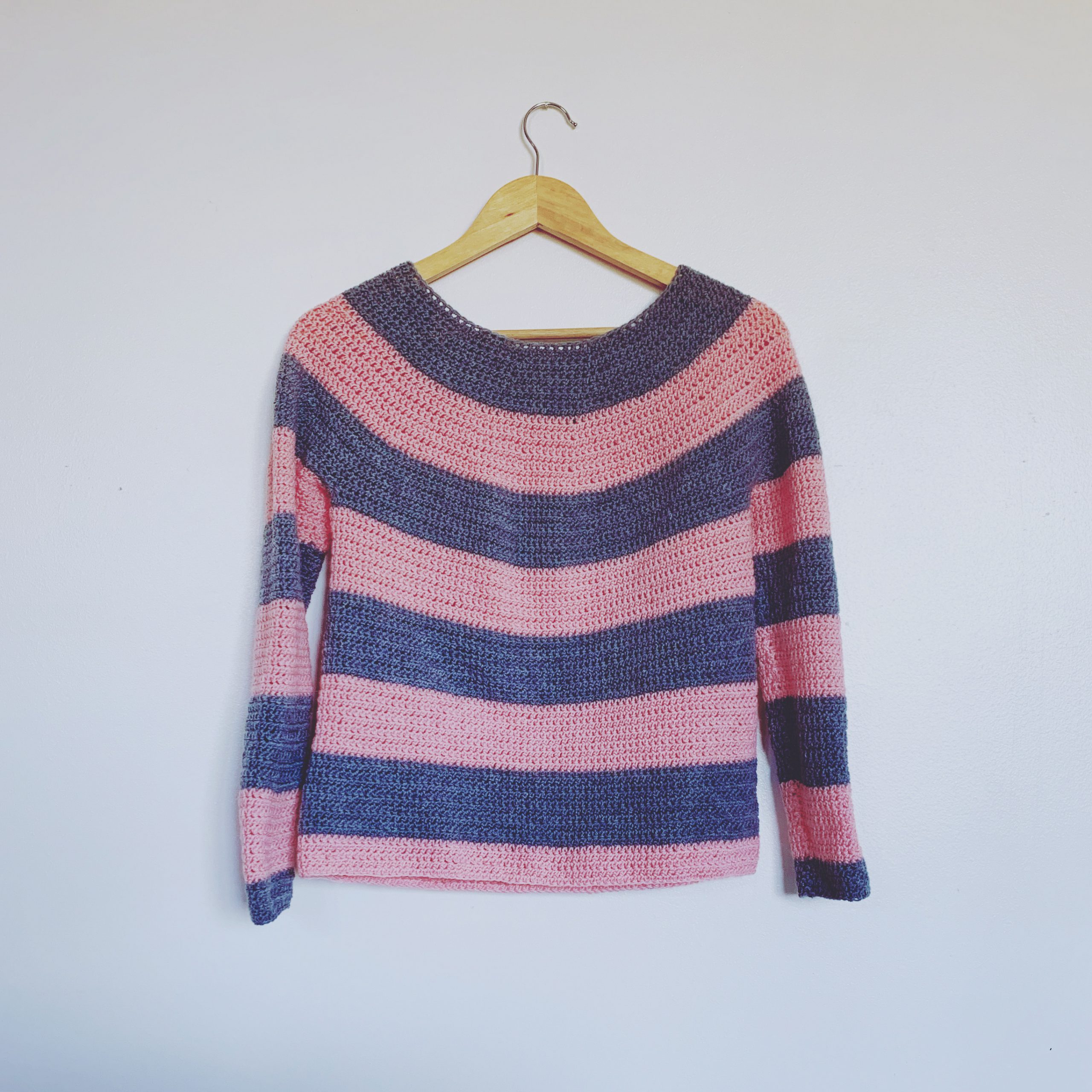 A 4ply pink and grey crochet striped sweater hangs on a white wall