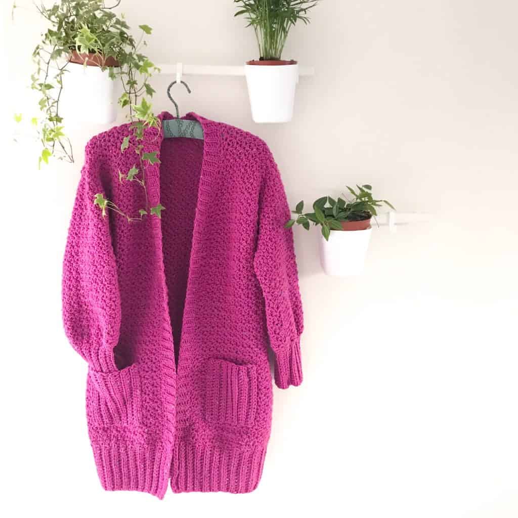 A long chunky pink crochet cardigan with ribbed pockets hangs on a white wall surrounded by plants
