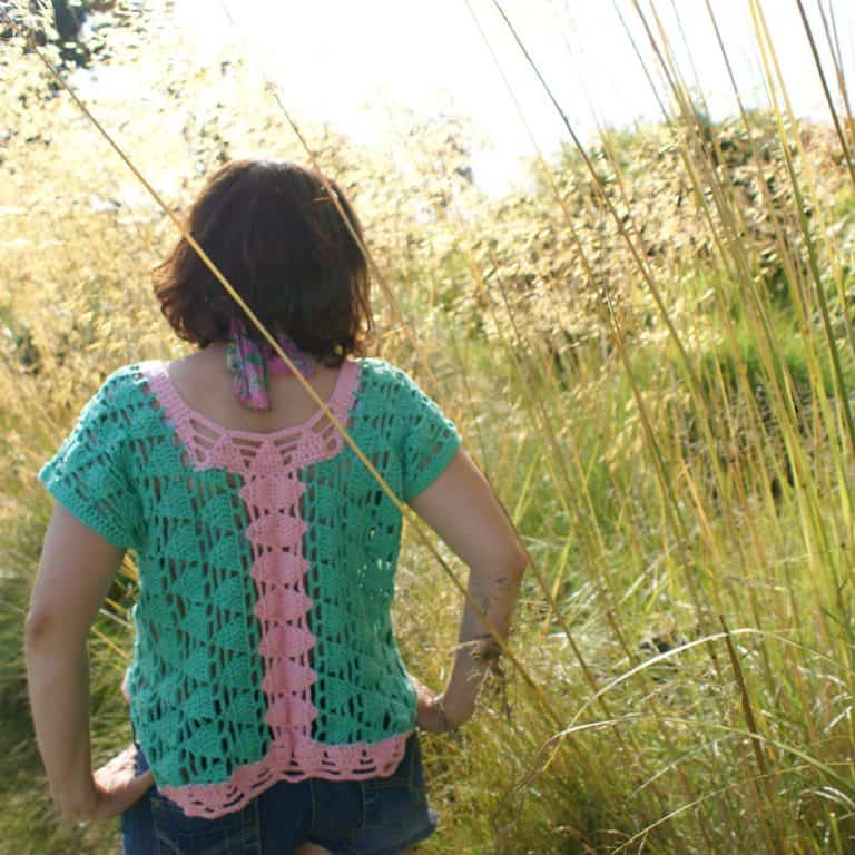 A woman stands back to camera with her hands on hips in a grass land wearing a pink and green triangle motif crochet top