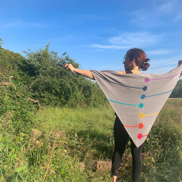 A woman stands with her back to the camera on a meadow holding out a chakra inspired crochet shawl
