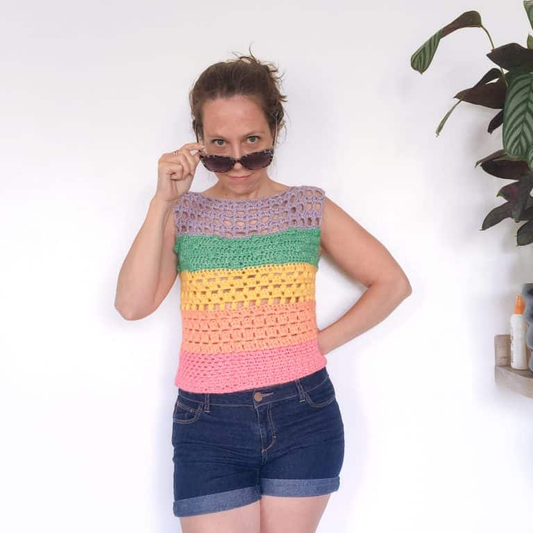 a woman in a bright crochet top and denim short stands in front of a white wall looking at the camera over her sunglasses