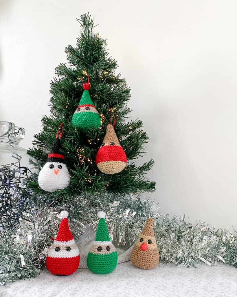 3 crochet Christmas decorations, including a Santa elf and rudolph gather round the bottom of a small christmas tree whilst a snowman elf and robin hang in the tree