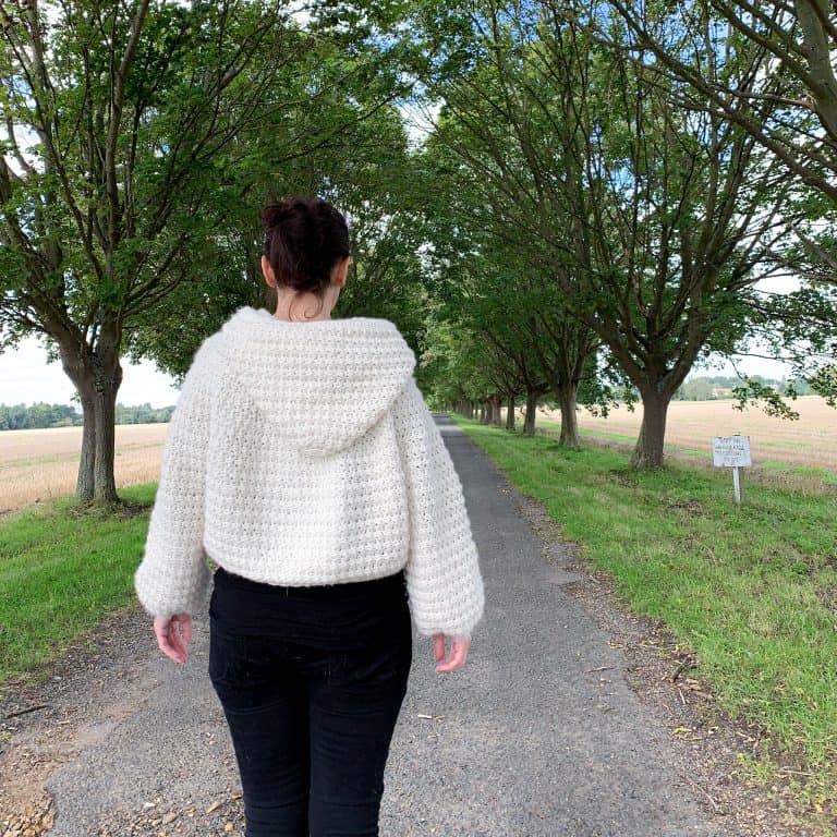 A woman with her back to the camera wearing black jeans and a natural wool crochet hooded cape stands in the centre of a tree lined avenue with fields to either side. Her hands fall by her side