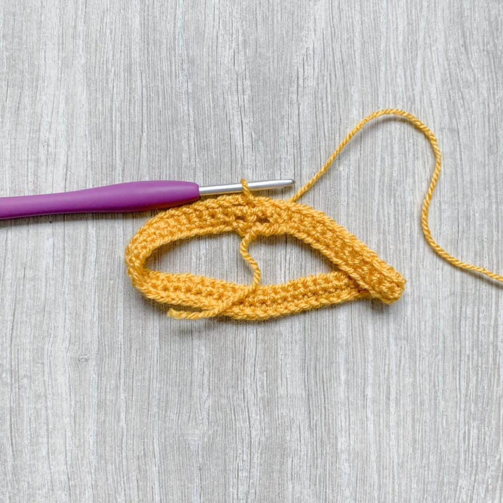 A tutorial step to make a crochet bow shows three rounds or crochet in yellow stitches with a purple crochet hook still looped in the last stitch