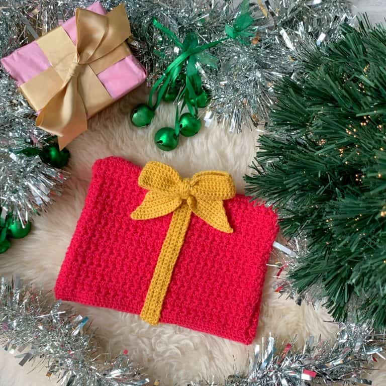 a pink crochet gift shaped christmas hat, with yellow ribbon and bow, lies on a sheepskin rug under a Christmas tree surrounded with green bells, tinsel and a gift wrapped in pink wrapping with a gold bow