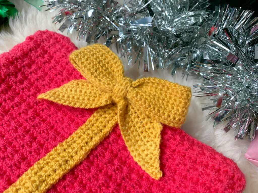 A yellow crochet bow is attached to a pink crochet rectangle surrounded by tinsel, with branches of a christmas tree encroaching