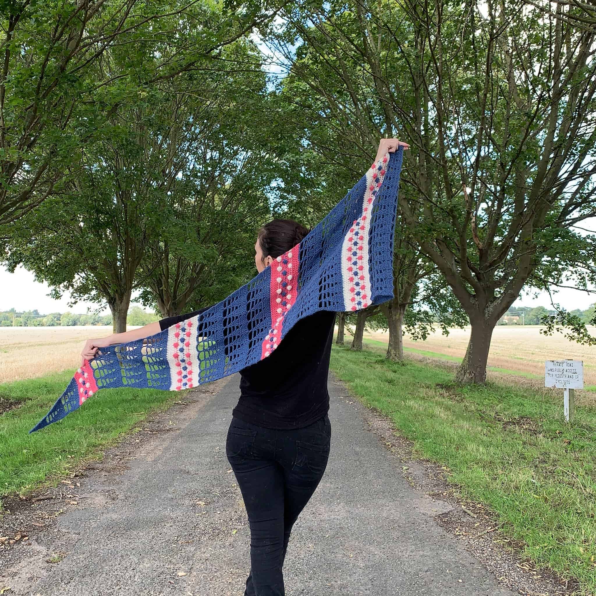 A woman stands in the centre of a tree lined avenue wearing all black. Her arms are outstretched holding either end of a blue, cream and pink crochet shawl which is flapping in the breeze. Her back is to the camera