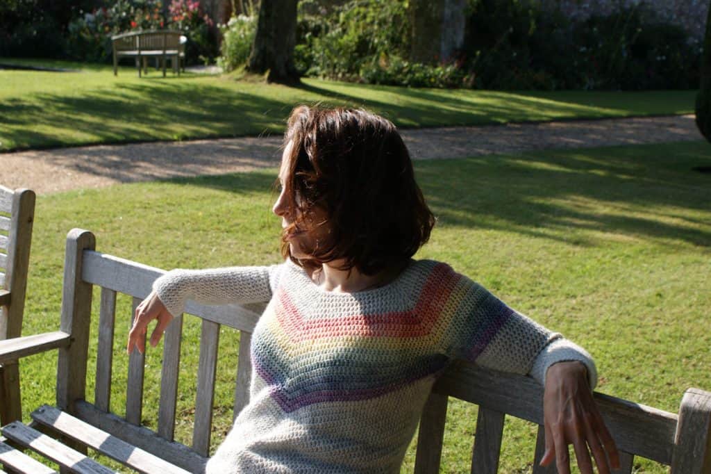 Dora wears the rainbow smiles crochet sweater whilst sitting on a bench in a lush garden late afternoon sun with her arms draped over the back