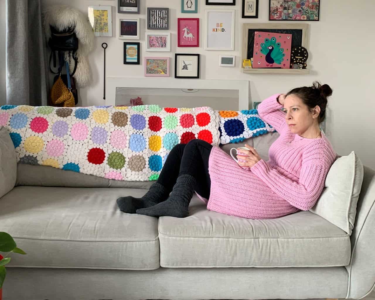 Dora sits on her sofa wearing a pink version of the winter solsitice crochet dress. She holds a cup of tea on her lap and looks out the window