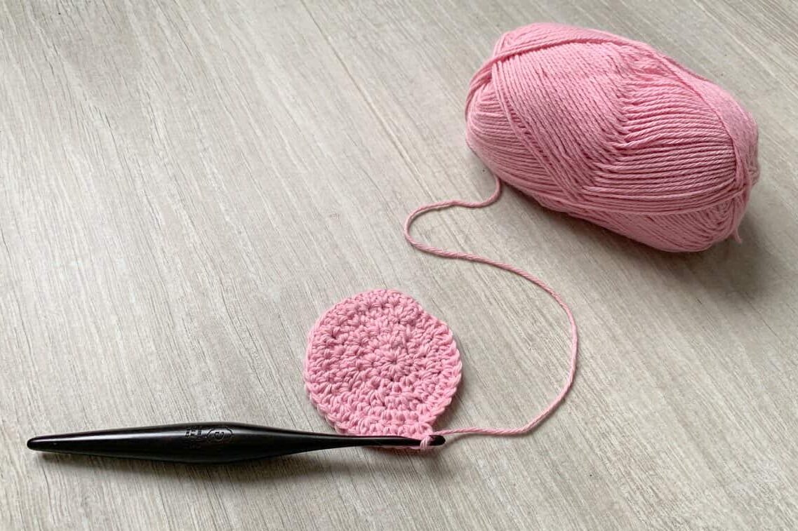 A small crochet circle with the last round partially worked is still attached to it’s pink ball of yarn with a black hook attached