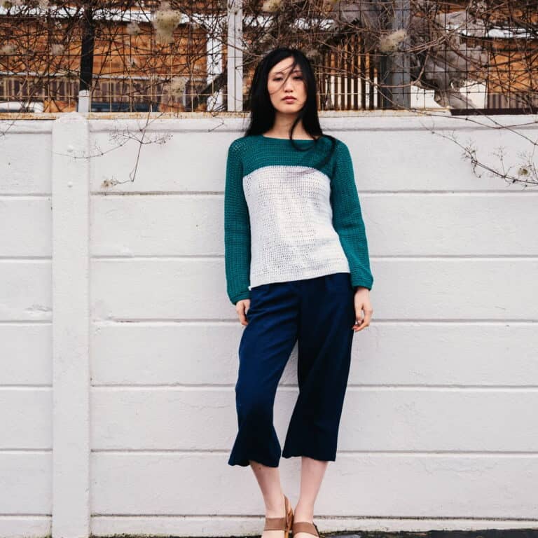 A woman stands in front of a white wall wearing the clarity crochet sweater, a teal and green jumper made with linen and cotton yarn