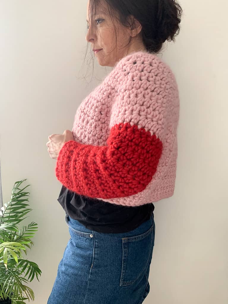 A woman wears a super chunky crochet cardigan with a pink body and clashing red sleeves