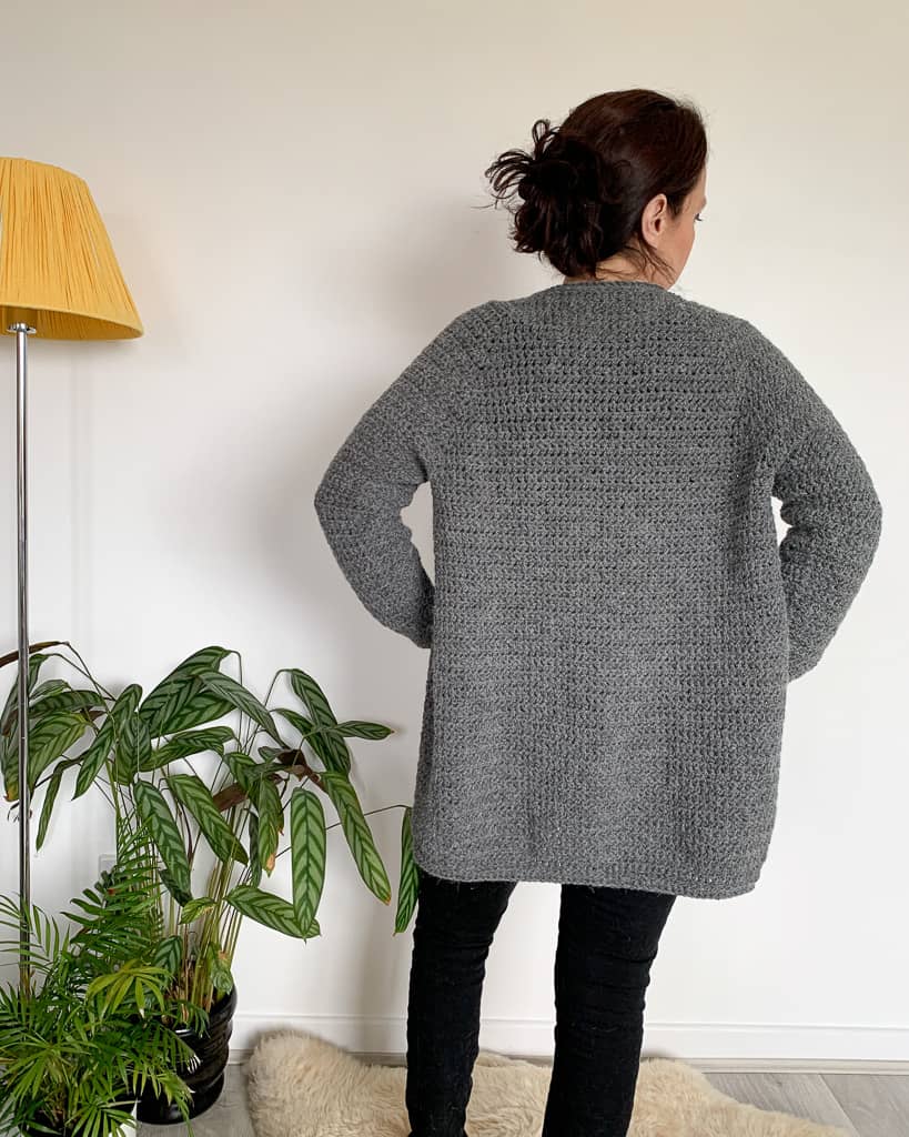 The back view of a woman wearing a long grey crochet cardigan, standing with her hands on hips.