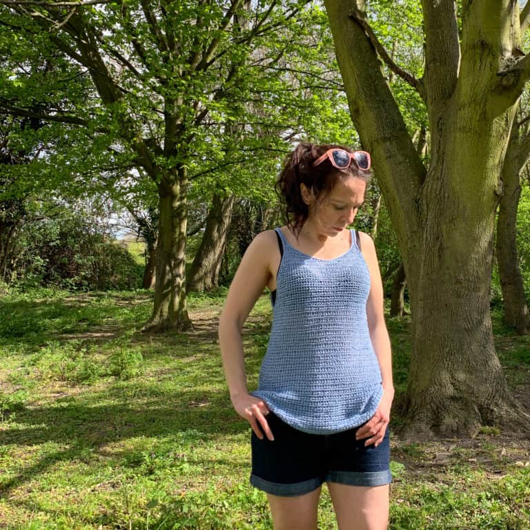 Dora hooks her thumbs in the pockets of her denim shorts as she looks to the floor of a wooded area, modelling a blue floaty crochet vest top on a sunny spot.