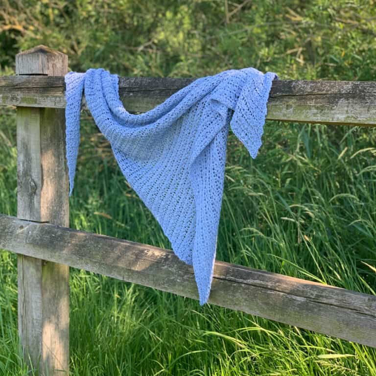 a blue crochet triangle shawl made with star stitch hangs from a wooden fence in a grassy meadow.