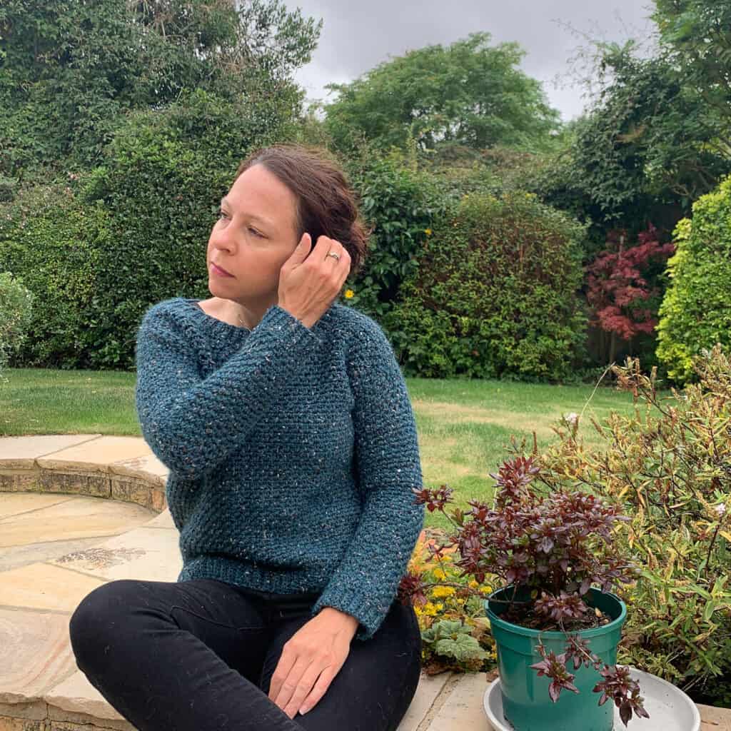 Dora is sat on a garden wall tucking her hair behind an ear whilst modelling the upsidedown crochet sweater pattern made in peacock blue tweed wool yarn.