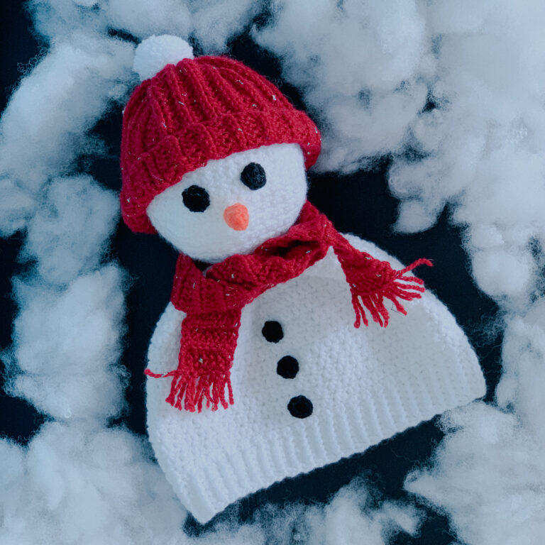 An adult size crochet snowman hat lies flat on a black background surrounded by fake snow.