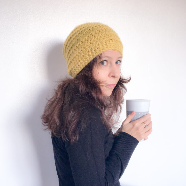 A woman holding a coffee cup and wearing a yellow crochet beanie looks into the camera