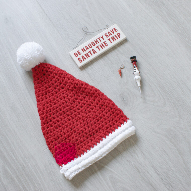 A red crochet Santa hat lies on a grey surface with a sign next to it reading ‘be naughty, save santa the trip'