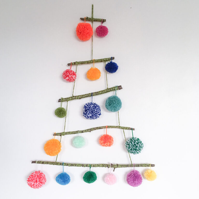 A ‘pom pom tree’ made of sticks tied tighter with pom poms hanging off them, hangs on a white wall.