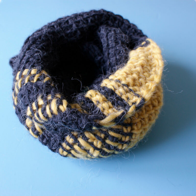 A yellow an charcoal grey Tunisian crochet scarf curled in a loop on a blue background.