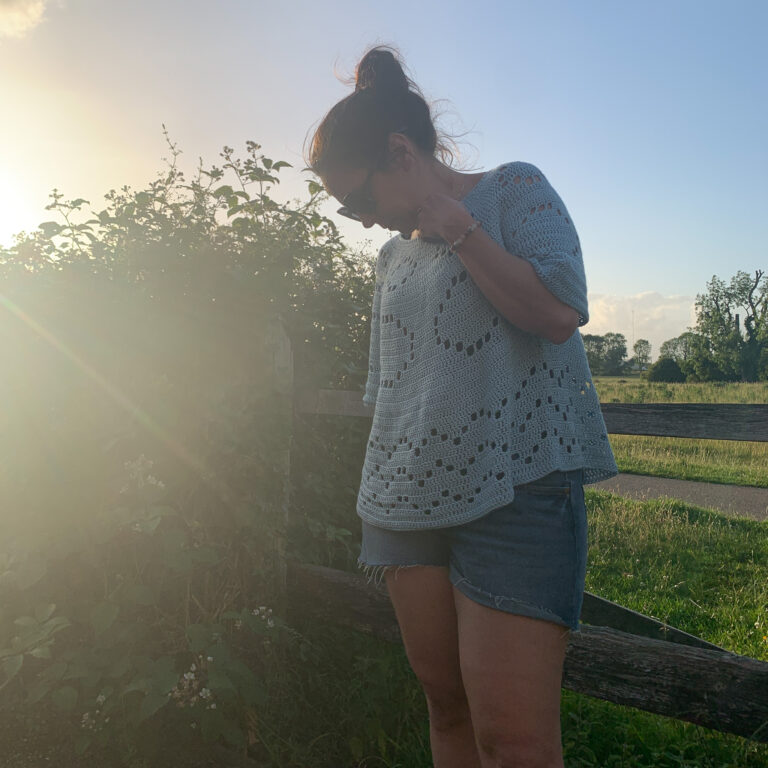 A woman wearing a blue crochet oversized short sleeve sweater with denim shorts and sunglasses looks to the ground to avoid the glare of the sunset.