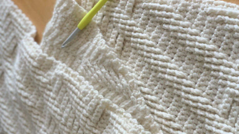 A close up of a cream textured crochet wrap with a yellow crochet hook on top.