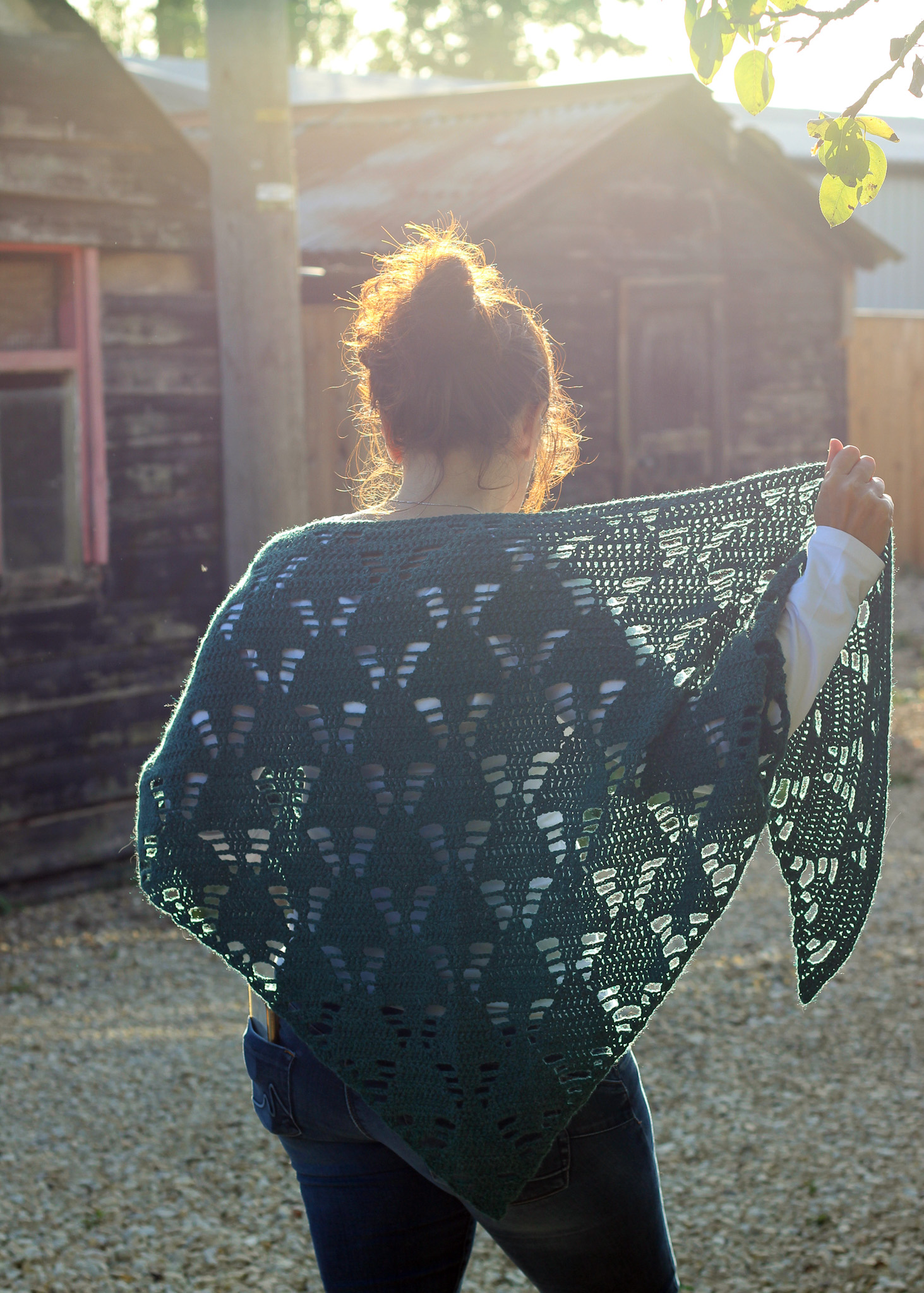 Dora is seen from behind wearing the under the tree crochet shawl with one side held away from her shoulder.