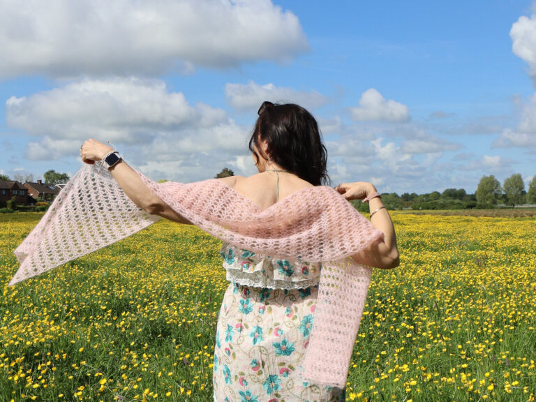 Dora stands in a field of buttercups wearing a pink mohair crochet wrap, with a summer dress. She holds the wrap out to the side so it catches the wind.