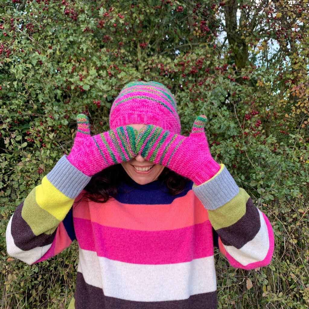 Dora hides her eyes between bright pink and green striped crochet mittens whilst wearing a matching crochet beanie hat.