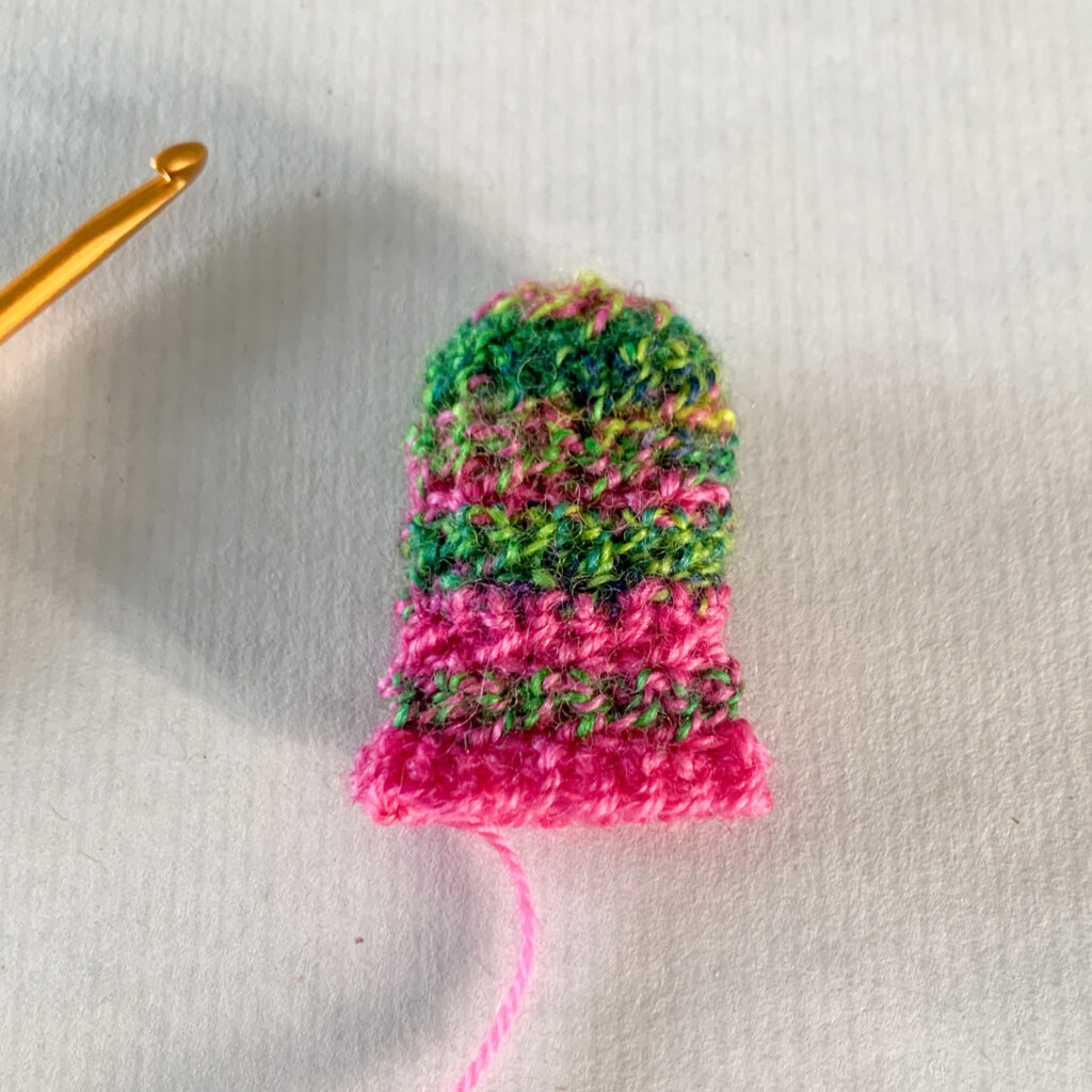 The thumb of a crochet mitten with stripes of pink and green yarn.