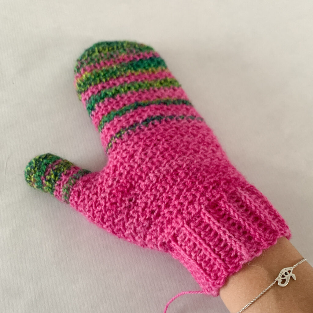 A hand resting on a white background wearing a pink crocheted mitten with torrent stripes over the fingers and thumb