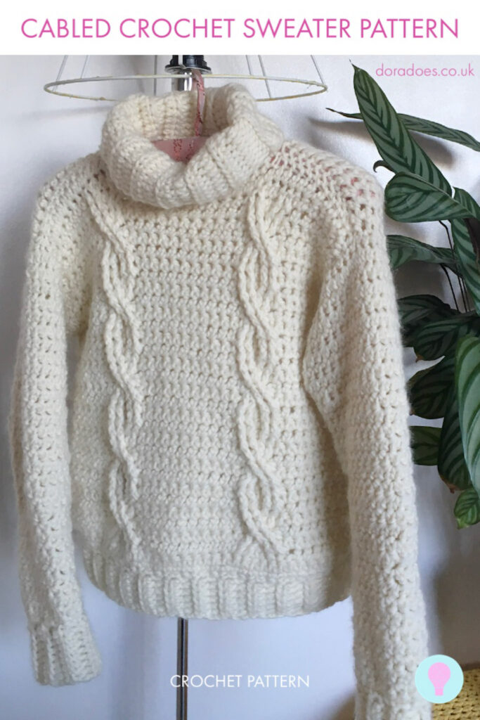 A cream cabled chunky crochet sweater with a roll neck and two parallel twisted cables down the front hangs from a lampshade. A plant is behind it and a text overlay reads ‘cabled crochet sweater pattern.’