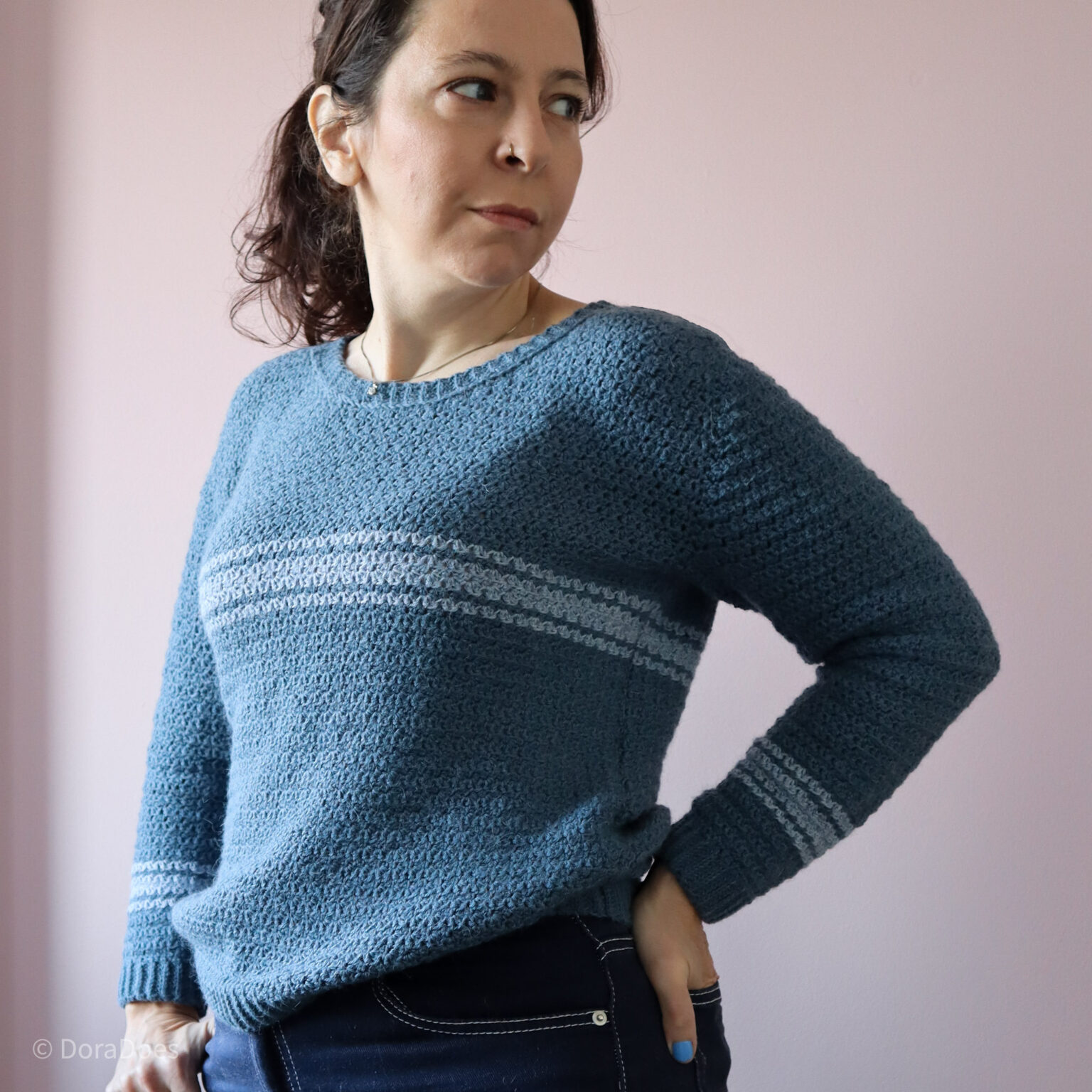 A woman wearing a blue crochet sweater with pale stripes across the chest and long sleeves stands looking to the side, hand resting on hip.