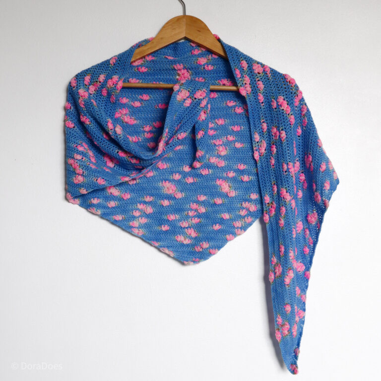 A blue crochet shawl with pink puff stitch bobbles is draped over a hanger with one end tucked in showing the drape and movement of the crocheted wrap.