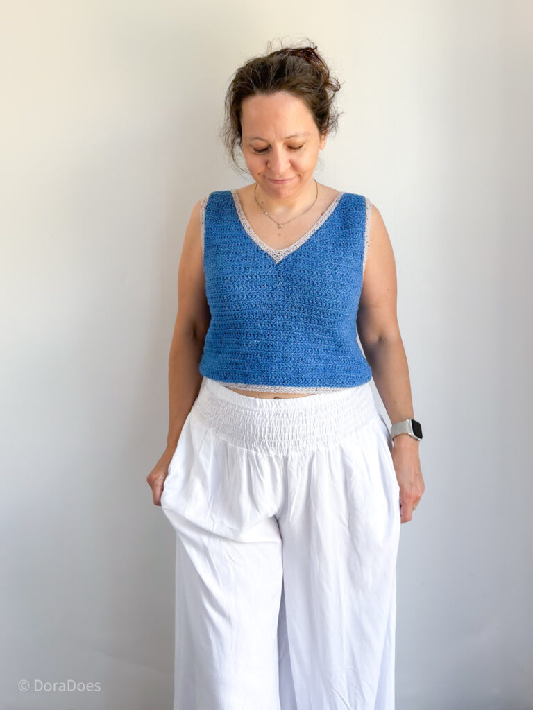 Dora stands in front of a white wall wearing a blue v-neck crochet tank top paired with wide leg white trousers. She is smiling as she lifts up one of the legs of the trousers.
