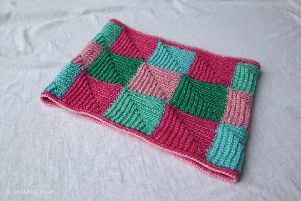 A crochet cowl made from square motifs lies flat on a white sheet.