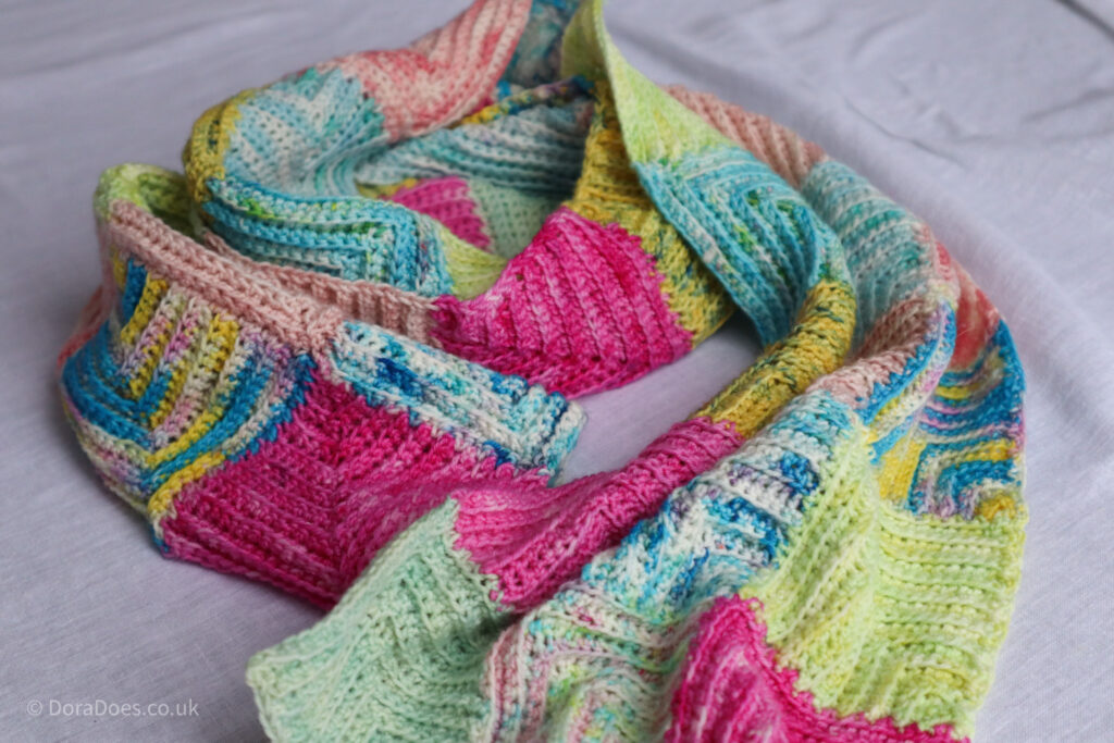 A crochet scarf with a patchwork look lies coiled on a white sheet.