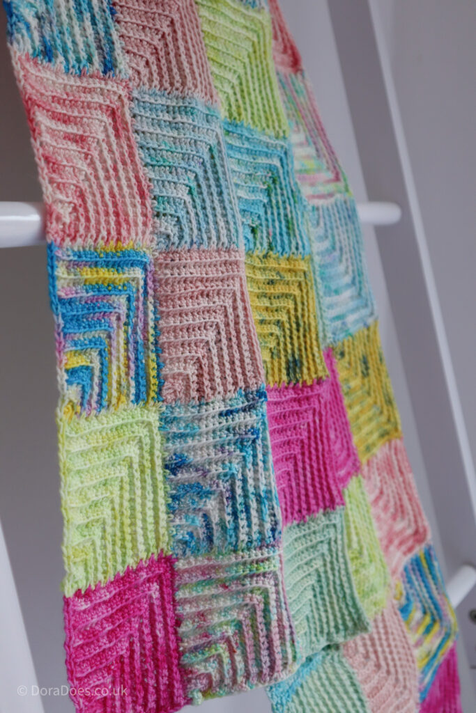 A close up of a crochet scarf handing on a rail, made from mitred squares in a range of hand dyed yarns.
