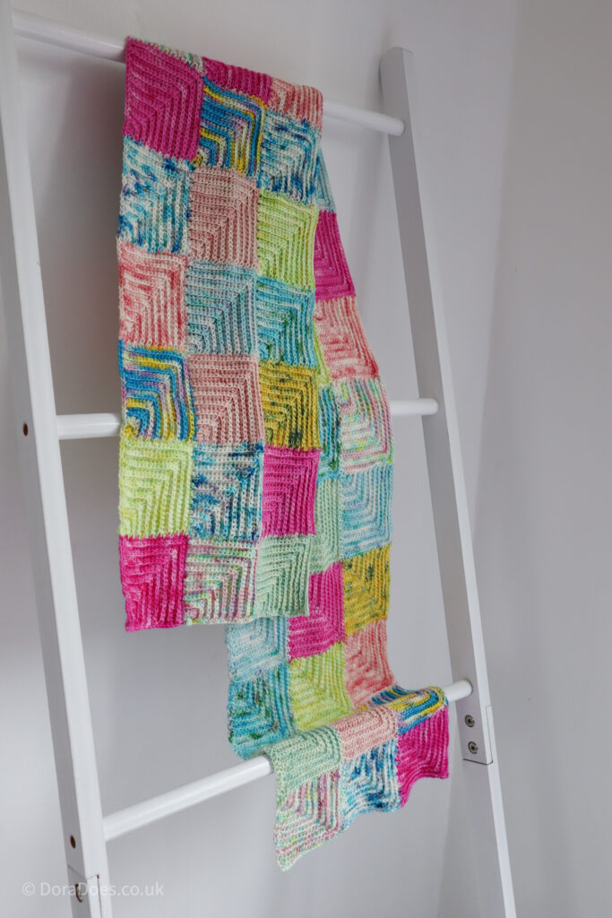 A patchwork crochet scarf made from mitered squares hangs over a blanket ladder.
