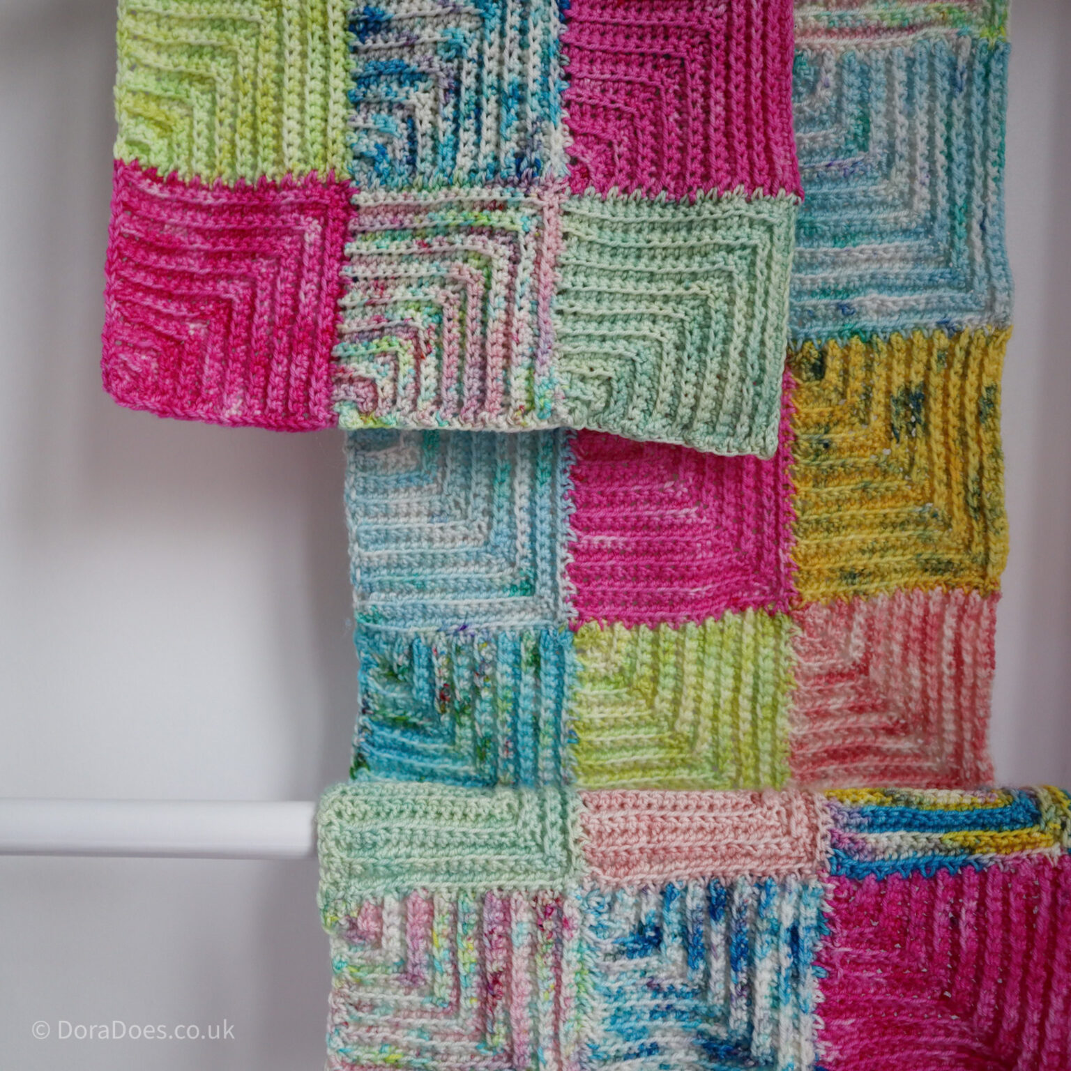 A square image of a crochet scarf made from mitered squares in a patchwork of hand dyed yarns hanging and overlapping itself on a blanket ladder.