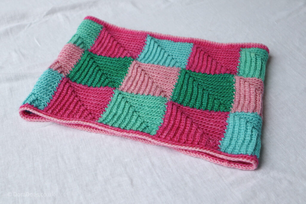 A crochet cowl made from joined crochet mitered squares in shades of pinks and greens lies on a white sheet.