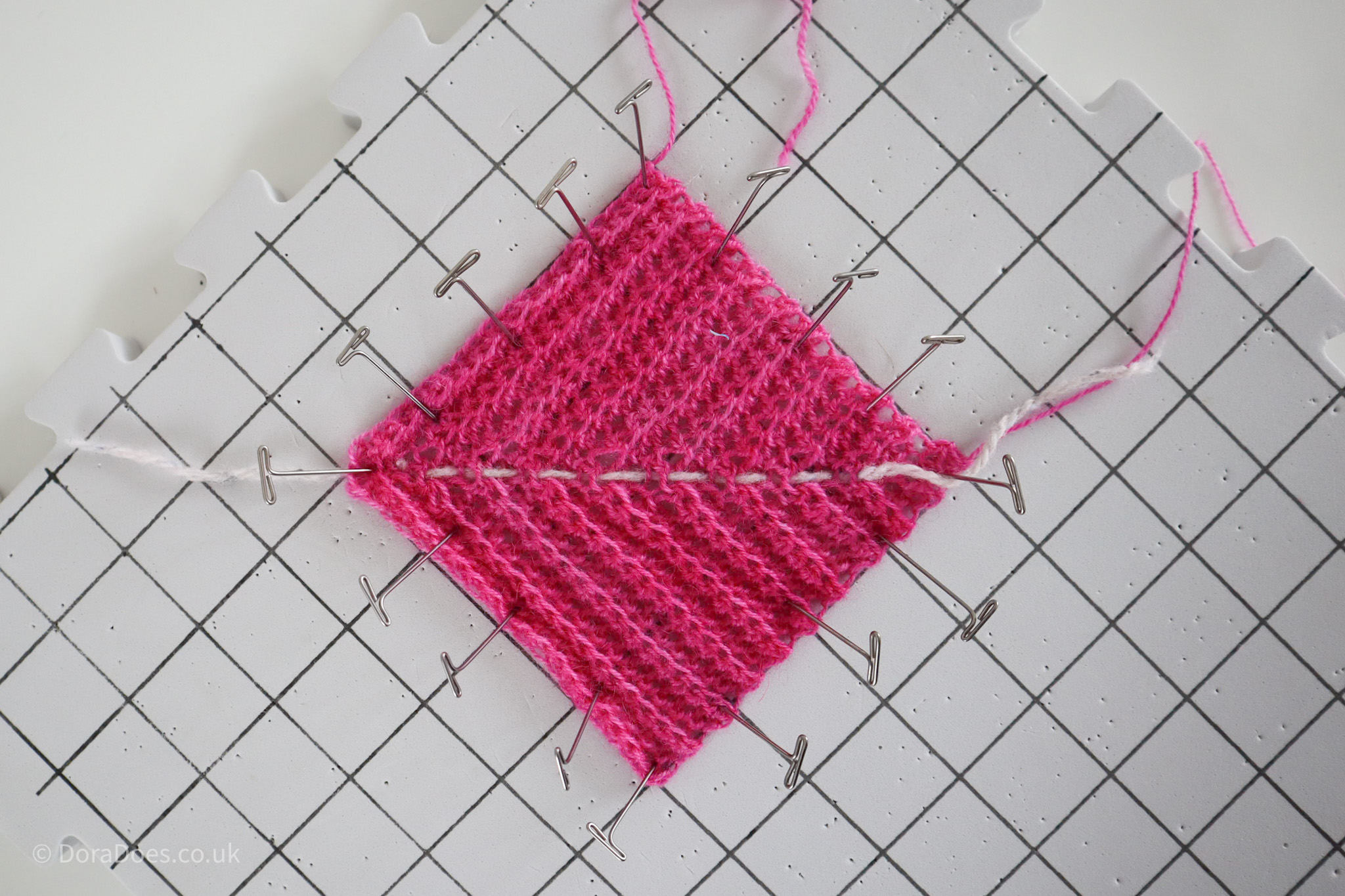 A mitred square in pink yarn is seen from above pinned to a blocking board.