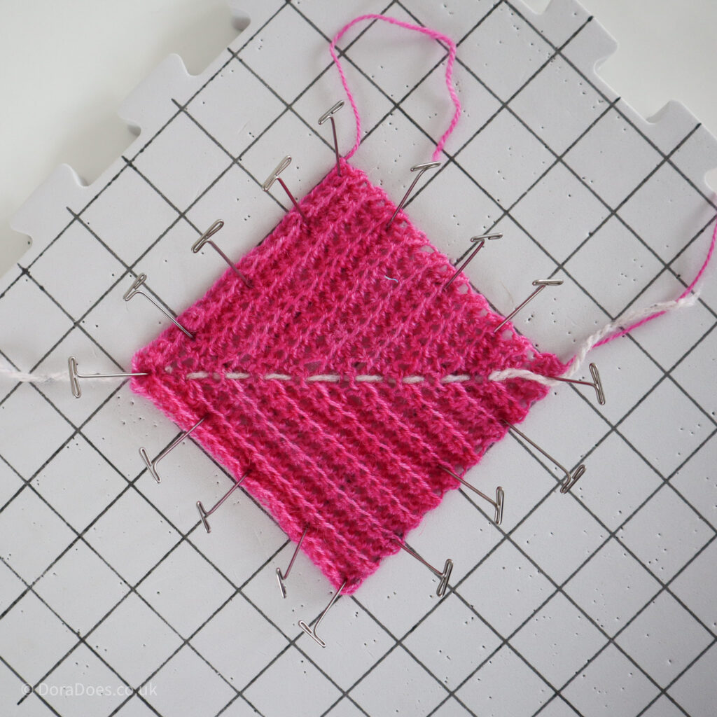 A pink crochet mitred square with ribbon stitch is pinned to a blocking board and seen on the diagonal.
