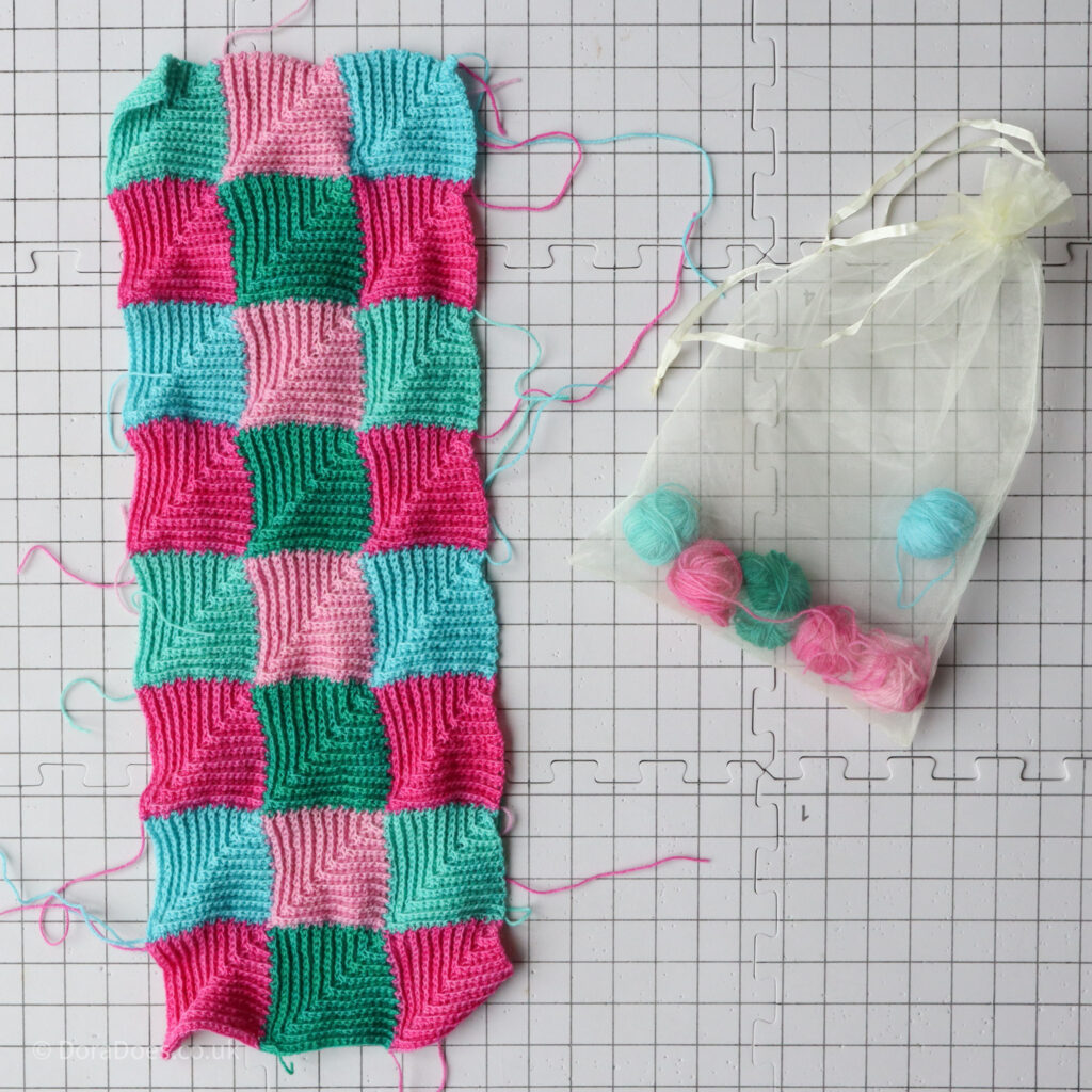 A series of joined crochet mitred squares lays flat on a blocking board with a clear bag of the leftover yarn placed next to it.