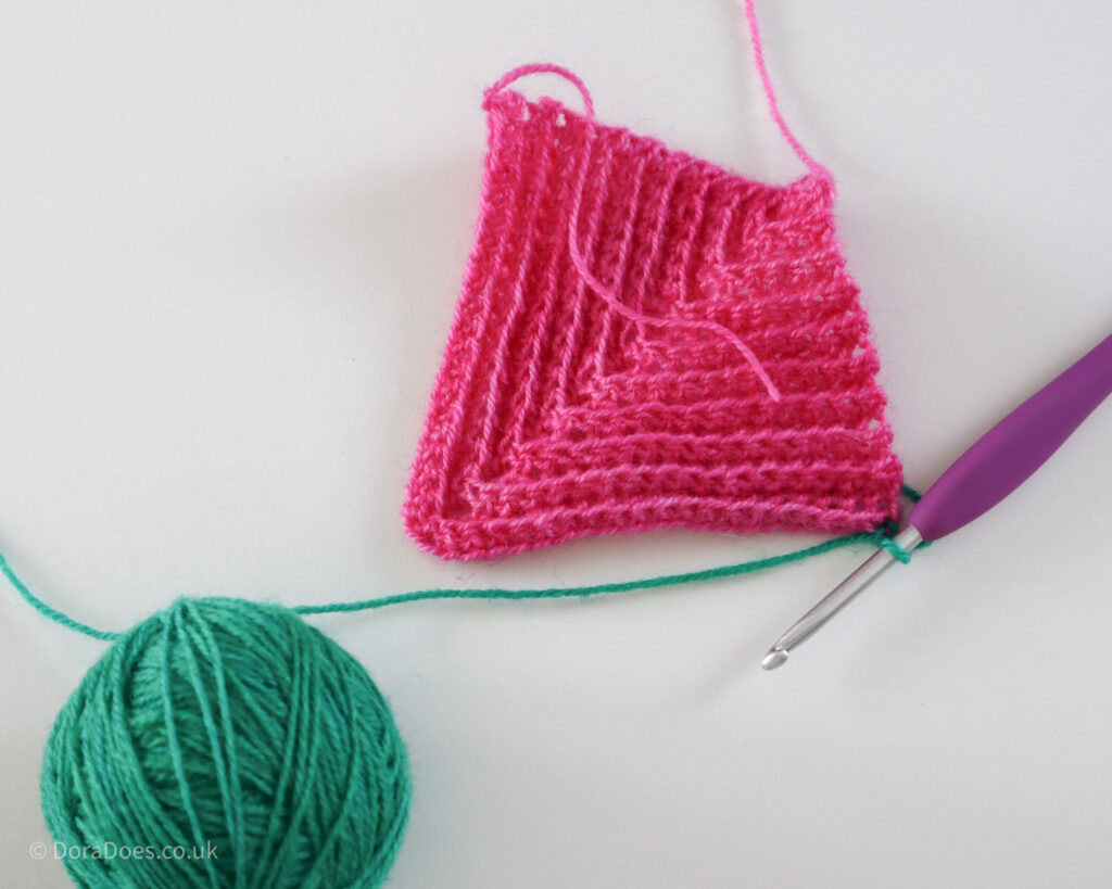 A crochet hook joins a ball of green yarn to a completed pink crochet mitred square.